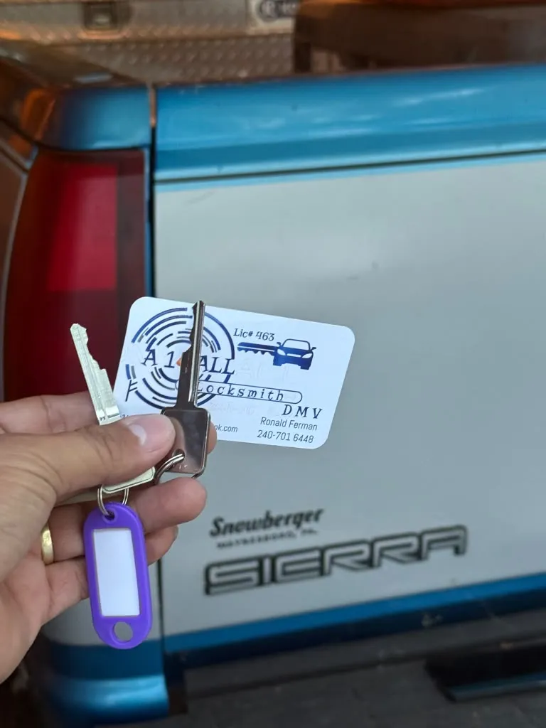 Two cut vehicle keys on a ring with a purple tag and an A1 All Locksmith DMV business card held at the tailgate of a GMC Sierra pickup